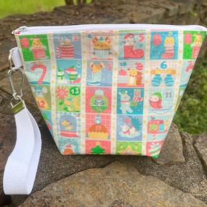 May include: A zippered pouch featuring a colorful grid pattern of holiday-themed illustrations, including animals, ornaments, and numbered squares from one to twenty-four. The bag includes a white fabric wrist strap attached with a silver metal clip.