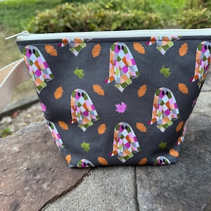 May include: A dark gray zippered pouch featuring a repeating pattern of ghosts with colorful checkered bodies and scattered orange and green autumn leaves. The bag includes a cream-colored fabric wrist strap attached to the side and a white zipper.