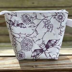 May include: A grey zippered pouch with a white zipper and a white handle. The pouch has a floral and bat print in shades of grey and black.