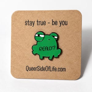 Puede incluir: Un pin de esmalte verde con forma de rana con una expresión escéptica y el texto "REALLY?" en su cuerpo. El pin está sujeto a una tarjeta cuadrada de color tostado con el texto "stay true - be you" arriba y "QueerSideOfLife.com" abajo.