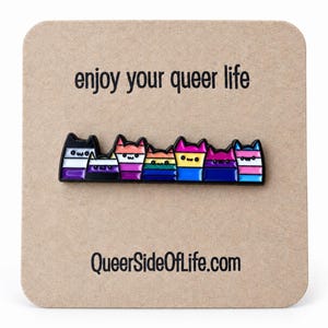 May include: A metal enamel pin featuring a row of seven stylized cats, each colored with different pride flag patterns. The pin is attached to a square tan card with the text "enjoy your queer life" at the top and "QueerSideOfLife.com" at the bottom.