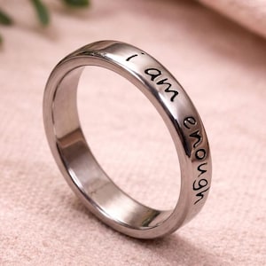 Peut inclure: Une bague en métal argenté avec une finition lisse et polie. La surface extérieure de l'anneau présente le texte gravé "i am enough" dans une police noire simple en minuscules. La bague est circulaire avec une largeur constante.
