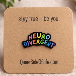 Peut inclure: Une broche en émail noir avec le texte NEURO DIVERGENT en lettres aux couleurs de l'arc-en-ciel. La broche est fixée sur une carte carrée beige avec le texte stay true - be you en haut et l'adresse QueerSideOfLife.com imprimée en bas.