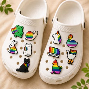 Könnte beinhalten: Ein Paar weiße Clogs, verziert mit verschiedenen bunten Pride-Anhängern, darunter ein Frosch, eine Schnecke, ein Hase, eine Katze, ein Einhorn, eine Ente und eine Gans. Die Anhänger zeigen Regenbogenmuster und den Schriftzug PURRIDE.