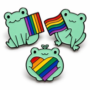 Puede incluir: Tres pines de esmalte con forma de ranas de color verde claro y contornos negros. Dos ranas sostienen banderas rectangulares con los colores del arcoíris, mientras que la tercera rana en el centro sostiene un gran corazón arcoíris.