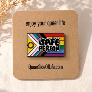 Puede incluir: Un pin esmaltado rectangular con el diseño de la bandera Progress Pride, que incluye un círculo amarillo y un anillo morado a la izquierda, y el texto negro "SAFE PERSON" con un pequeño corazón rosa a la derecha, sobre una tarjeta beige.