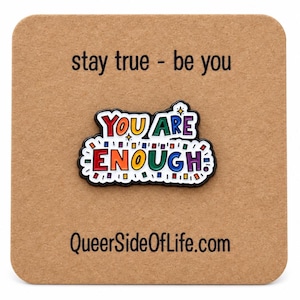 Peut inclure: Une broche en émail métallique avec le texte YOU ARE ENOUGH en lettres multicolores et des accents de confettis. La broche est fixée sur une carte carrée marron imprimée avec le texte stay true - be you et Queersideoflife.com.