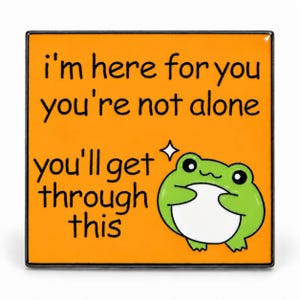 Op de afbeelding: Een vierkante oranje emaille pin met een zwarte rand en zwarte tekst: i'm here for you you're not alone you'll get through this. Een kleine groene kikker met een witte buik en een klein wit sterretje bevindt zich in de rechterbenedenhoek.