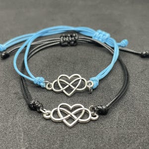 Pode incluir: Duas pulseiras de cordão ajustável com berloques de metal prateado em forma de coração infinito. Uma pulseira é feita com um cordão azul claro, enquanto a outra é feita com um cordão preto, ambas incluindo nós deslizantes para ajuste.