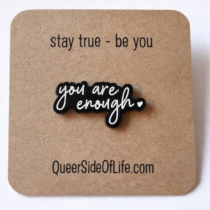 Może przedstawiać: Czarny emaliowany pin z białym napisem "you are enough" i małym sercem. Pin znajduje się na brązowej karcie z napisami "stay true - be you" i "QueerSideOfLife.com".