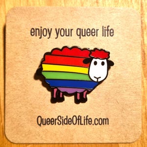 Könnte beinhalten: Ein bunter Emaille-Anstecker mit einem Schaf-Design, das einen Regenbogenkörper und schwarze Akzente aufweist. Der Anstecker befindet sich auf einem beigefarbenen Hintergrund mit dem Text "enjoy your queer life" und "QueerSideOfLife.com".