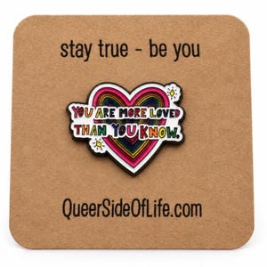 Op de afbeelding: Een hartvormige emaille speld met een regenboogkleurig centrum en de tekst YOU ARE MORE LOVED THAN YOU KNOW. De speld is bevestigd op een vierkante bruine kaart bedrukt met stay true - be you en QueerSideOfLife.com.