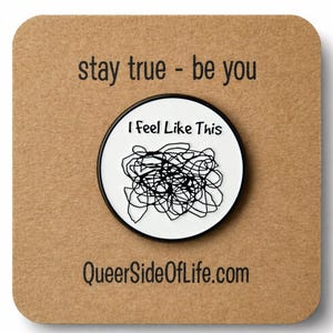 Pode incluir: Um pin redondo com borda preta e o texto "I Feel Like This" em fundo branco, com um desenho de rabiscos pretos. Está sobre um cartão castanho com os textos "stay true - be you" e "QueerSideOfLife.com".