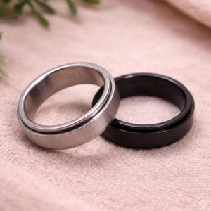 Könnte beinhalten: Zwei Spinner-Ringe, einer mit einer gebürsteten silberfarbenen Metalloberfläche und der andere mit einer glatten, mattschwarzen Oberfläche. Beide Ringe haben ein breites Banddesign mit einem drehbaren Mittelteil, das sich frei bewegen lässt.