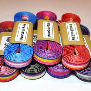 Può includere: Una collezione di lacci colorati, ciascuno arrotolato e fissato con una fascia di carta marrone. I lacci presentano una varietà di schemi di colori arcobaleno e sfumati, tra cui rosso, blu, rosa e viola. Il testo "Outer Side of Life" è stampato sulle fasce di carta.