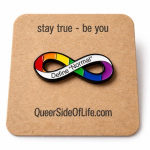 Könnte beinhalten: Eine unendlichkeitsförmige Emaille-Anstecknadel mit Regenbogenverlauf und dem schwarzen Text "Define "Normal"". Die Anstecknadel ist auf einer quadratischen beigen Karte befestigt, die oben mit "stay true - be you" und unten mit "QueerSideOfLife.com" bedruckt ist.