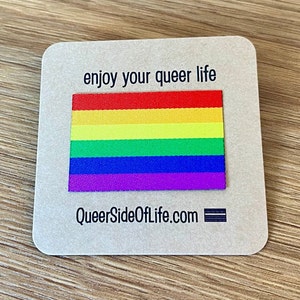 May include: A brown card with the text "enjoy your queer life" and a rainbow flag. The card also has the text "QueerSideOfLife.com" and a black and white stripe.