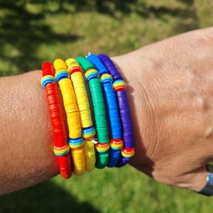 Puede incluir: Un conjunto de cinco pulseras apiladas hechas de cuentas planas y circulares de arcilla polimérica en un espectro arcoíris de rojo, amarillo, verde, azul y morado. Cada pulsera incluye pequeñas cuentas redondas con rayas.