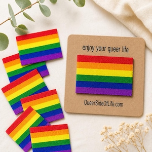 Könnte beinhalten: Rechteckige Stoffaufnäher mit einem horizontalen Regenbogenflaggen-Design in Rot, Orange, Gelb, Grün, Blau und Violett. Ein Aufnäher ist auf einer quadratischen beigen Karte befestigt, die mit dem Text enjoy your queer life und QueerSideOfLife.com bedruckt ist.