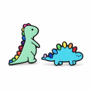 Puede incluir: Dos pines de esmalte con forma de dinosaurios de dibujos animados con contornos negros. Uno es un dinosaurio verde claro de pie con una espina dorsal de colores del arcoíris. El otro es un dinosaurio azul horizontal con placas dorsales de colores.