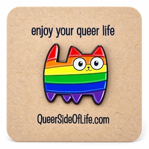 Peut inclure: Une broche émaillée en forme de chat aux couleurs de l'arc-en-ciel avec des contours noirs, de grands yeux blancs et un petit nez. La broche est fixée sur une carte carrée beige avec le texte "enjoy your queer life" et "QueerSideOfLife.com".