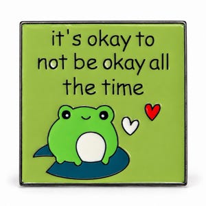 May include: A square enamel pin with a light green background featuring a green frog sitting on a dark blue lily pad. The text reads, "it's okay to not be okay all the time." A small white heart and a small red heart are positioned to the right.