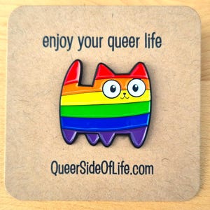 Op de afbeelding: Een kleurrijke katvormige emaille pin met een regenboogontwerp, met rode, oranje, gele, groene, blauwe en paarse strepen. De pin heeft een zwarte omtrek en grote witte ogen. De tekst op de kaart luidt "enjoy your queer life" en "QueerSideOfLife.com".
