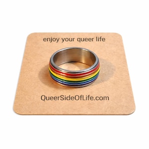 May include: A stainless steel ring with a rainbow-colored band, displayed on a brown card. The ring features red, orange, yellow, green, blue, and purple stripes. The card reads "enjoy your queer life" and "QueerSideOfLife.com".