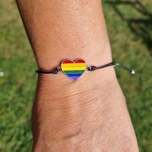 Könnte beinhalten: Ein schwarzes Kordelarmband mit einem herzförmigen Anhänger. Der Anhänger zeigt ein Regenbogen-Design mit roten, orangefarbenen, gelben, grünen, blauen und violetten Streifen. Das Armband wird am Handgelenk getragen.