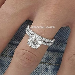 Può includere: Primo piano di una mano che indossa un anello di fidanzamento in argento con un grande diamante rotondo centrale e una fascia di diamanti più piccola. L'anello è incastonato a griffe e i diamanti brillano.