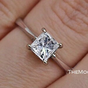May include: A silver ring with a square-shaped diamond set in a prong setting. The ring is on a finger.
