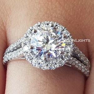 May include: A silver engagement ring with a large round center stone surrounded by a halo of smaller stones. The band is split into two bands with smaller stones set into the band.