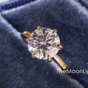 May include: A round, clear diamond engagement ring set in a gold band. The ring is displayed in a blue velvet ring box. TheMoonLights is printed on the box.