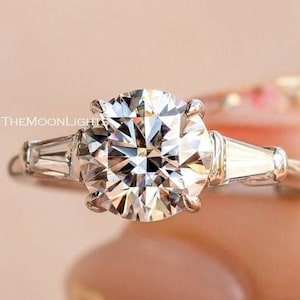 May include: A close-up of a diamond engagement ring with a large, round center stone and two smaller baguette-cut diamonds on either side. The ring is set in a silver band. The text "THEMOONLIGHT" is visible.