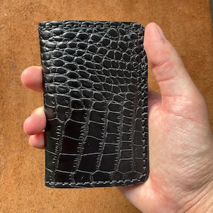 Puede incluir: Cartera de cuero negro texturizado con un patrón de piel de cocodrilo. La cartera rectangular parece cosida a mano y tiene un acabado brillante. Se sostiene en una mano. Fondo marrón claro.