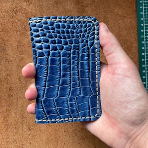 May include: A blue leather card holder with a crocodile skin texture. The edges are stitched with a light beige thread. The card holder is rectangular and appears to be new.
