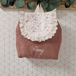 Puede incluir: Una mochila de pana color rosa empolvado con una solapa blanca de estampado floral delicado y ribete de encaje de ojal color crema. El nombre Romy est&aacute; bordado en letra cursiva blanca en la parte frontal de la bolsa, que incluye un asa.