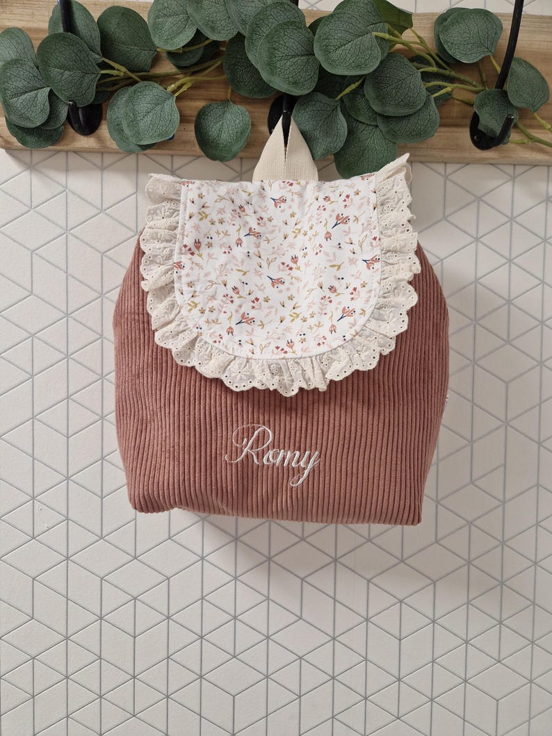 Puede incluir: Una mochila de pana color rosa empolvado con una solapa blanca de estampado floral delicado y ribete de encaje de ojal color crema. El nombre Romy est&aacute; bordado en letra cursiva blanca en la parte frontal de la bolsa, que incluye un asa.