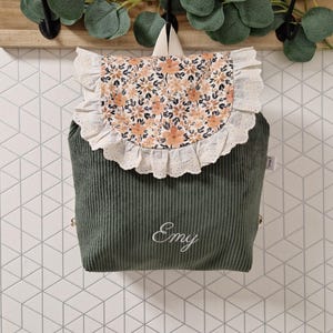 Puede incluir: Una mochila de pana verde salvia con una solapa de estampado floral y un borde de encaje blanco con volantes. La parte frontal tiene bordado el nombre Emy en letras cursivas blancas. La mochila incluye un asa superior y correas para los hombros.