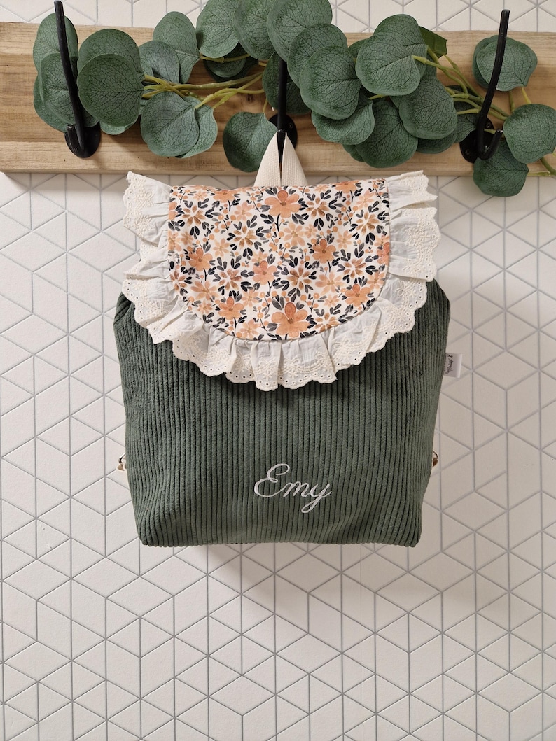 Puede incluir: Una mochila de pana verde salvia con una solapa de estampado floral y un borde de encaje blanco con volantes. La parte frontal tiene bordado el nombre Emy en letras cursivas blancas. La mochila incluye un asa superior y correas para los hombros.