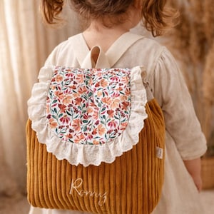 Puede incluir: Una mochila de pana color amarillo mostaza con una solapa redondeada que presenta un estampado floral colorido y un borde de encaje blanco. El nombre Romy est&aacute; bordado en blanco en la parte frontal, con correas de tela a juego incluidas.