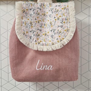 Puede incluir: Una mochila de pana rosa polvoriento con una solapa redondeada que presenta un estampado floral en blanco, amarillo y gris, adem&aacute;s de un borde con flecos color crema. El nombre Lina est&aacute; impreso en letra cursiva blanca en el frente de la bolsa.