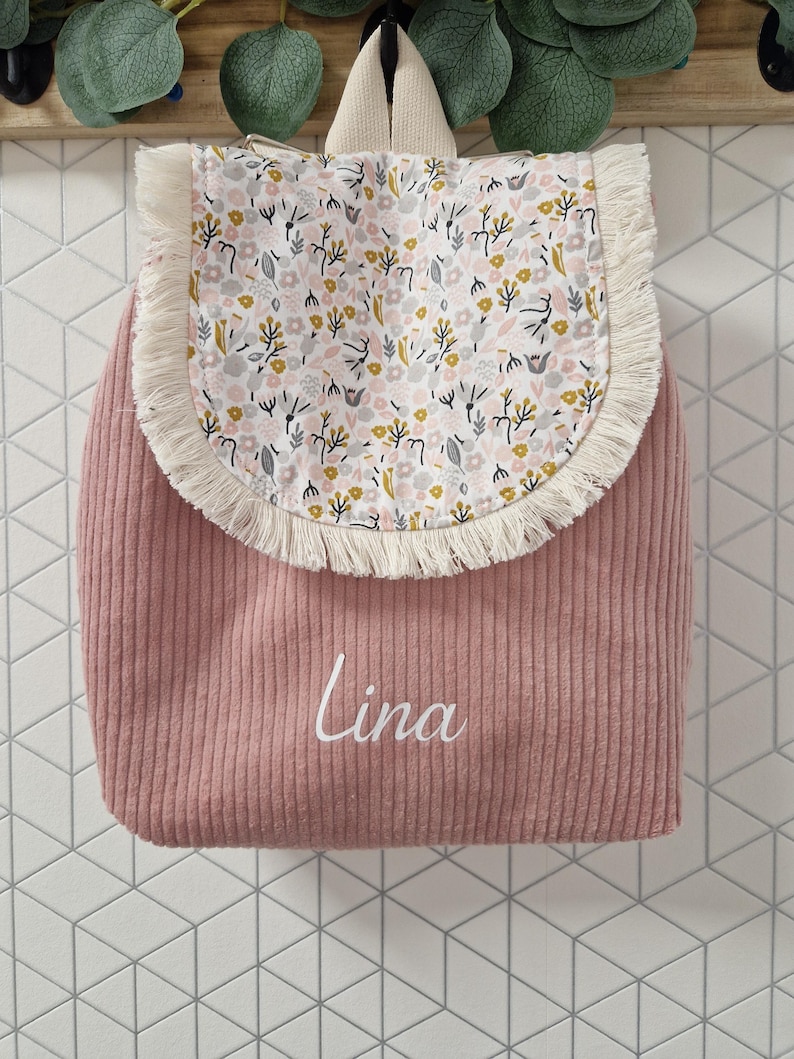 Puede incluir: Una mochila de pana rosa polvoriento con una solapa redondeada que presenta un estampado floral en blanco, amarillo y gris, adem&aacute;s de un borde con flecos color crema. El nombre Lina est&aacute; impreso en letra cursiva blanca en el frente de la bolsa.
