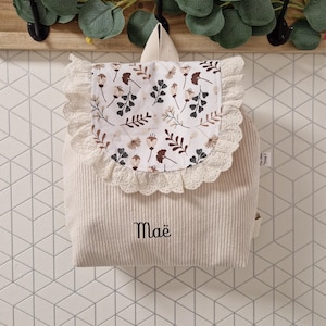 Puede incluir: Una mochila de pana color crema con una solapa que presenta un estampado floral en tonos marrones y negros. La solapa est&aacute; bordeada con encaje blanco. El nombre Ma&euml; est&aacute; bordado con hilo negro en la parte frontal de la mochila, que tiene asa.