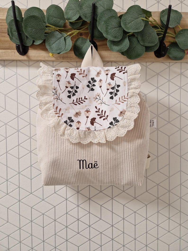 Puede incluir: Una mochila de pana color crema con una solapa que presenta un estampado floral en tonos marrones y negros. La solapa est&aacute; bordeada con encaje blanco. El nombre Ma&euml; est&aacute; bordado con hilo negro en la parte frontal de la mochila, que tiene asa.