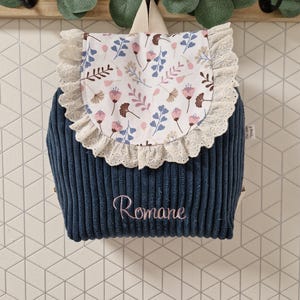 Puede incluir: Una mochila de pana azul marino con una solapa blanca con un estampado floral en rosa, azul y marr&oacute;n. La solapa est&aacute; adornada con encaje blanco. El nombre Romane est&aacute; bordado con hilo rosa en la parte delantera de la mochila.