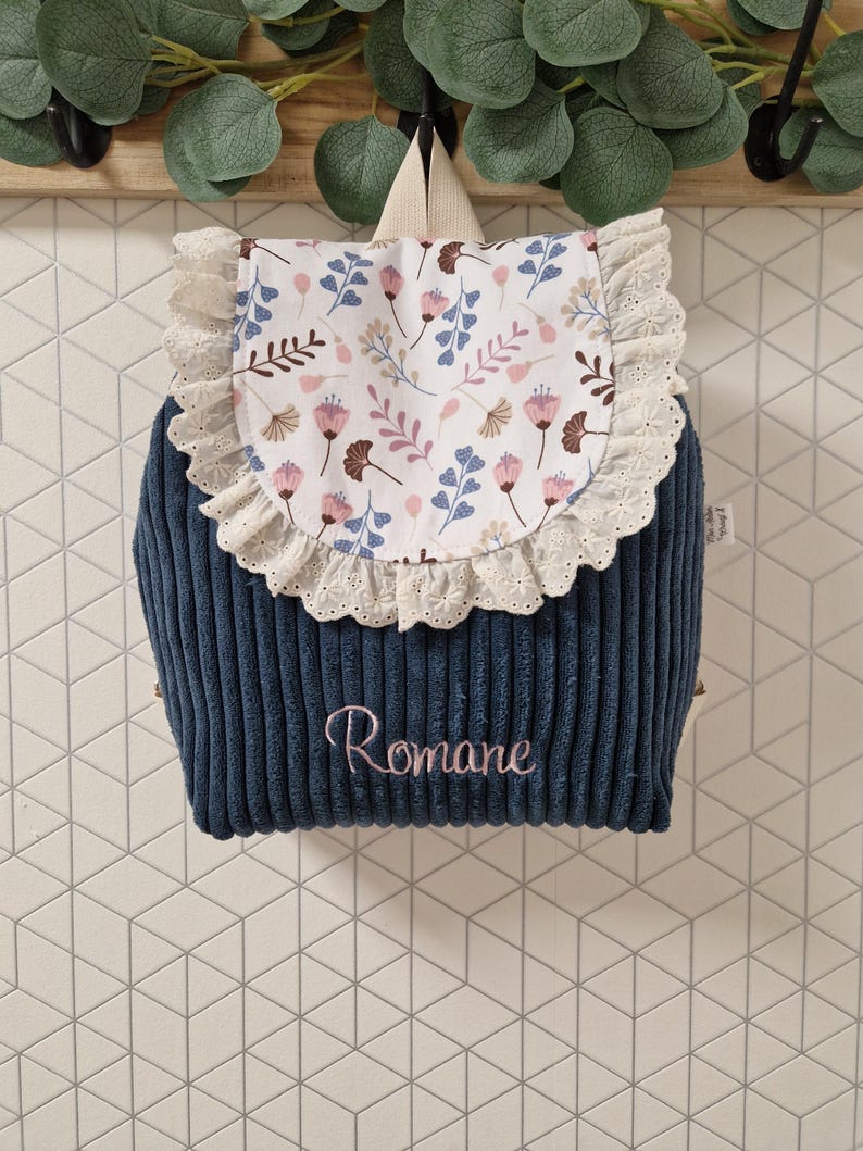 Puede incluir: Una mochila de pana azul marino con una solapa blanca con un estampado floral en rosa, azul y marr&oacute;n. La solapa est&aacute; adornada con encaje blanco. El nombre Romane est&aacute; bordado con hilo rosa en la parte delantera de la mochila.