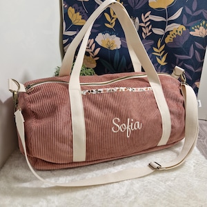 Peut inclure: Un sac polochon en velours côtelé rose poudré avec des poignées et une bandoulière crème. Le sac présente un motif floral et le nom "Sofia" brodé en cursive. Le sac est posé sur une surface blanche et moelleuse.