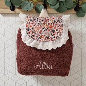 Peut inclure: Un sac à dos bordeaux avec un rabat à imprimé floral et une bordure en dentelle blanche. Le rabat présente un motif floral coloré avec des fleurs roses, oranges et blanches. Le nom "Alba" est brodé en écriture blanche.