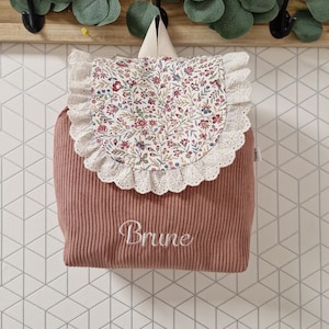 Puede incluir: Una mochila de pana color rosa empolvado con una solapa de estampado floral y ribete de encaje blanco. El nombre Brune est&aacute; bordado en cursiva blanca en la parte delantera. La mochila incluye un asa superior y una peque&ntilde;a etiqueta lateral.